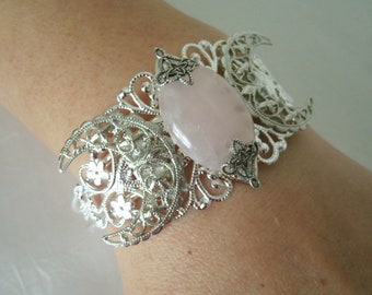 May include: A silver bracelet with a delicate filigree design featuring three crescent moons. A large, oval-shaped rose quartz stone is set in the center of the bracelet.