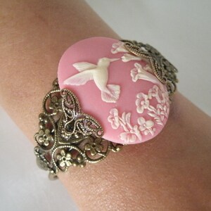 May include: A pink cameo bracelet with a white hummingbird and floral design. The bracelet is made of metal with an ornate filigree design.