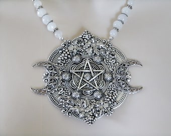 Puede incluir: Collar con colgante de plata con un pentagrama, lunas crecientes y diseños florales intrincados. El collar está enhebrado con cuentas blancas.