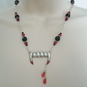 May include: A silver necklace with black and red beads and a silver pendant shaped like a set of vampire fangs with red accents. The pendant has a dangling charm with red beads.