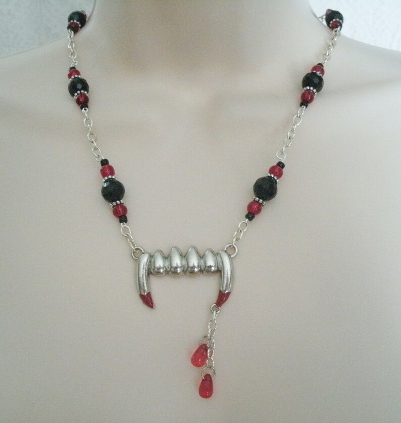 May include: A silver necklace with black and red beads and a silver pendant shaped like a set of vampire fangs with red accents. The pendant has a dangling charm with red beads.