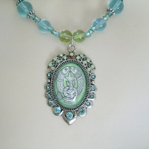 May include: A silver-toned necklace featuring a pendant with a light green background and a design of two fish. The pendant is surrounded by small blue gemstones and a beaded chain with blue and green beads.