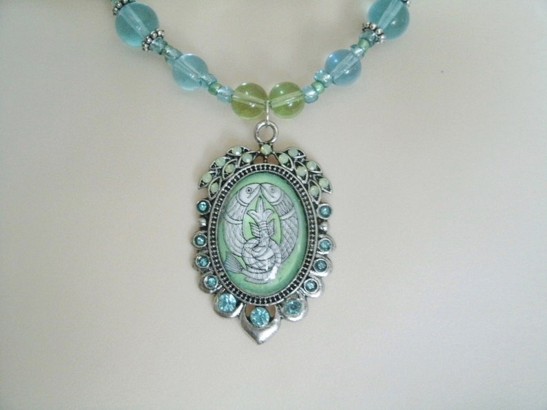 May include: A silver-toned necklace featuring a pendant with a light green background and a design of two fish. The pendant is surrounded by small blue gemstones and a beaded chain with blue and green beads.