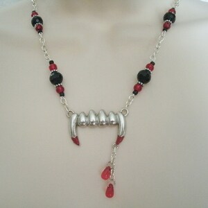 May include: A silver vampire fangs necklace with black and red beads. The necklace has a silver charm of fangs with red accents and a dangling charm of three red beads.