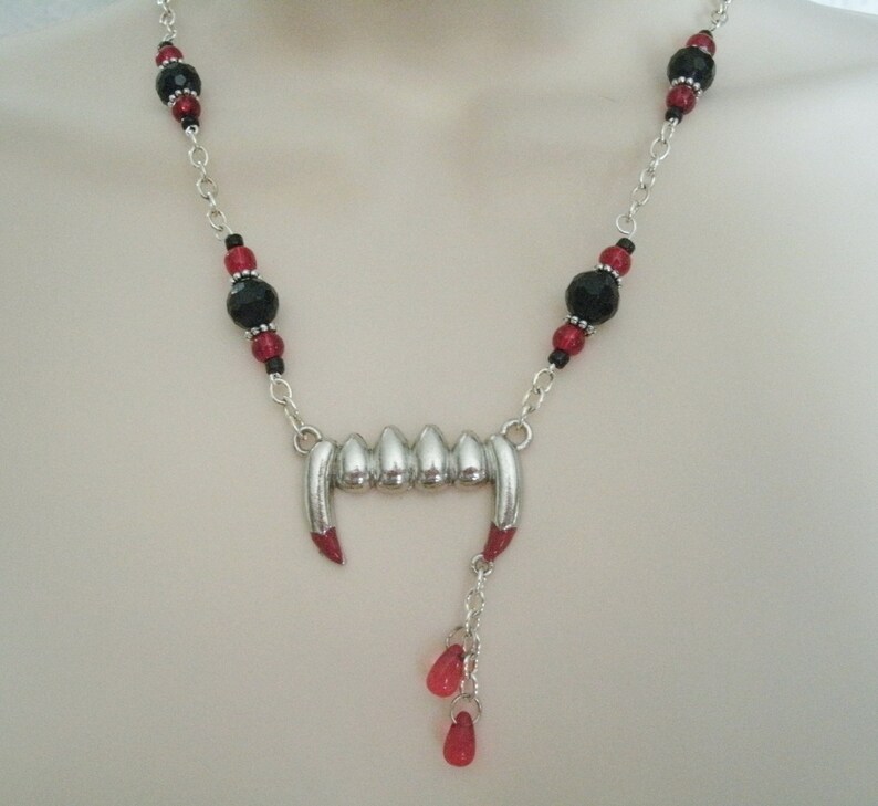 May include: A silver vampire fangs necklace with black and red beads. The necklace has a silver charm of fangs with red accents and a dangling charm of three red beads.