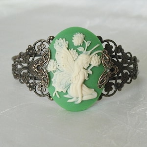 May include: A green cameo bracelet with a white cameo of a fairy with wings. The bracelet is made of metal with a filigree design.
