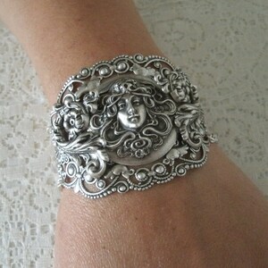 K&ouml;nnte beinhalten: Silberarmband mit einem verzierten Design, das ein Frauen Gesicht zeigt, umgeben von floralen Motiven.