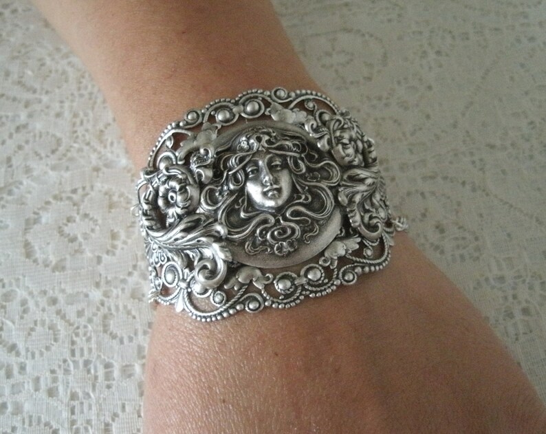 K&ouml;nnte beinhalten: Silberarmband mit einem verzierten Design, das ein Frauen Gesicht zeigt, umgeben von floralen Motiven.