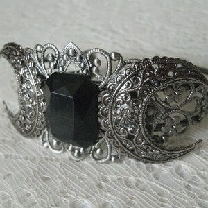 May include: A silver bracelet with a black gemstone in the center and two crescent moon designs on either side.