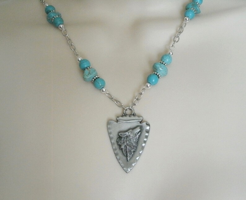May include: A silver necklace with a turquoise bead chain and a silver arrowhead pendant with a wolf head design.