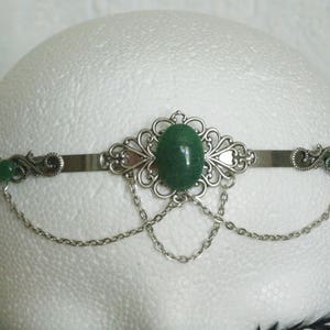 May include: Silver headband with a green gemstone and dangling chains. The headband is decorated with ornate silver filigree.