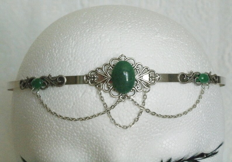 May include: Silver headband with a green gemstone and dangling chains. The headband is decorated with ornate silver filigree.