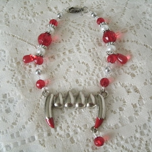 May include: A silver vampire fang necklace with red and white beads. The necklace is made of silver and has a silver clasp.