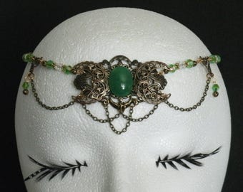 Puede incluir: Un tocado con una piedra ovalada verde central engastada en un marco de metal color bronce adornado con motivos de lunas crecientes. El diseño incluye delicadas cadenas de bronce y pequeñas cuentas verdes y transparentes en los laterales.