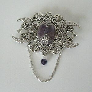 May include: A silver filigree brooch with a purple stone in the center and a dangling purple bead on a silver chain.