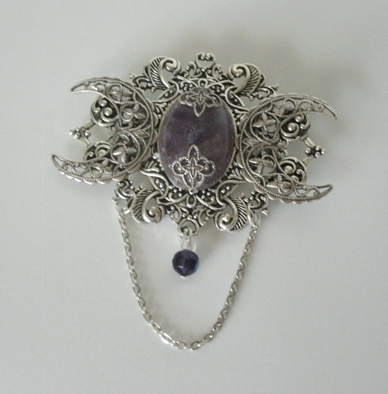 May include: A silver filigree brooch with a purple stone in the center and a dangling purple bead on a silver chain.