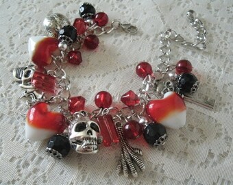 May include: A silver charm bracelet with red and black glass beads, a silver skull charm, a silver key charm, and a silver heart charm. The bracelet is on a white lace background.