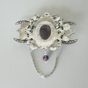 May include: Silver tone brooch with a large, ornate, filigree design. The brooch features a large, oval-shaped, dark purple stone in the center. The brooch has a chain with a small, iridescent bead hanging from it.
