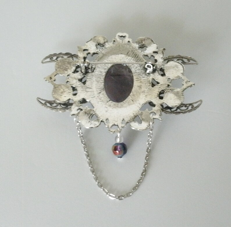 May include: Silver tone brooch with a large, ornate, filigree design. The brooch features a large, oval-shaped, dark purple stone in the center. The brooch has a chain with a small, iridescent bead hanging from it.