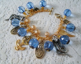 May include: A gold-toned charm bracelet featuring blue faceted beads and various charms. Charms include a cobra, a pentagram, a crescent moon, and a pair of wings. The bracelet is on a white lace surface.