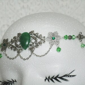 Puede incluir: Una diadema plateada y verde con una gran piedra preciosa verde, piedras preciosas verdes m&aacute;s peque&ntilde;as y dise&ntilde;os florales plateados.