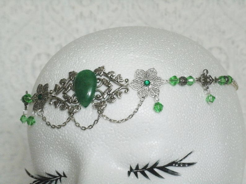 Puede incluir: Una diadema plateada y verde con una gran piedra preciosa verde, piedras preciosas verdes m&aacute;s peque&ntilde;as y dise&ntilde;os florales plateados.