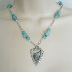 May include: A silver necklace with a turquoise bead chain and a silver pendant shaped like an arrowhead with a wolf's head inside.