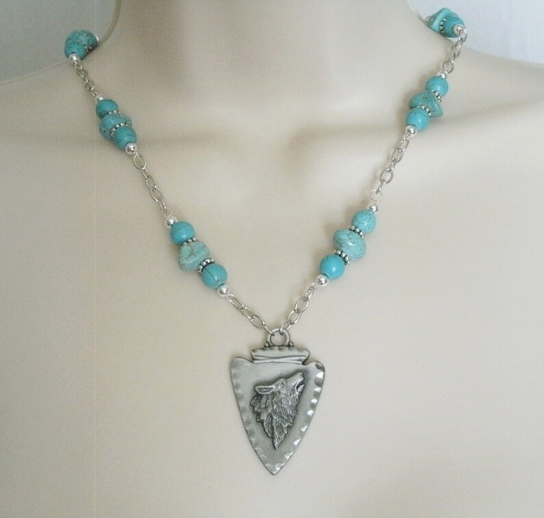 May include: A silver necklace with a turquoise bead chain and a silver pendant shaped like an arrowhead with a wolf's head inside.