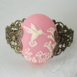 May include: A pink cameo bracelet with a white hummingbird and floral design. The bracelet is made of metal with a filigree design.