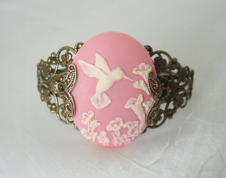 May include: A pink cameo bracelet with a white hummingbird and floral design. The bracelet is made of metal with a filigree design.
