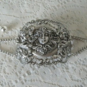 K&ouml;nnte beinhalten: Silberarmband mit einem detaillierten, verzierten Design, das ein Frauen Gesicht zeigt, umgeben von floralen Motiven.