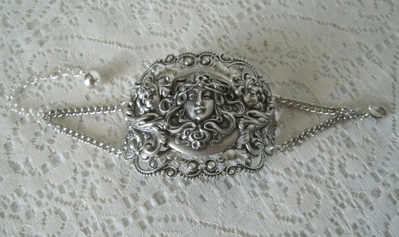 K&ouml;nnte beinhalten: Silberarmband mit einem detaillierten, verzierten Design, das ein Frauen Gesicht zeigt, umgeben von floralen Motiven.