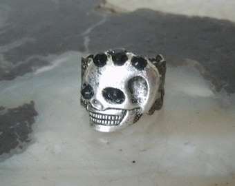 Puede incluir: Un anillo de calavera de plata con detalles de gemas negras. La calavera tiene un diseño detallado y está montada en una banda de plata.