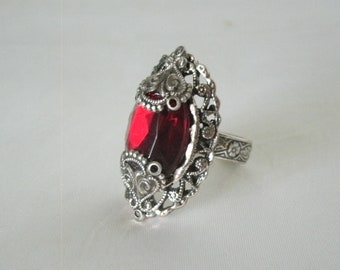 May include: A silver ring with an ornate design featuring a large, red, faceted gemstone.