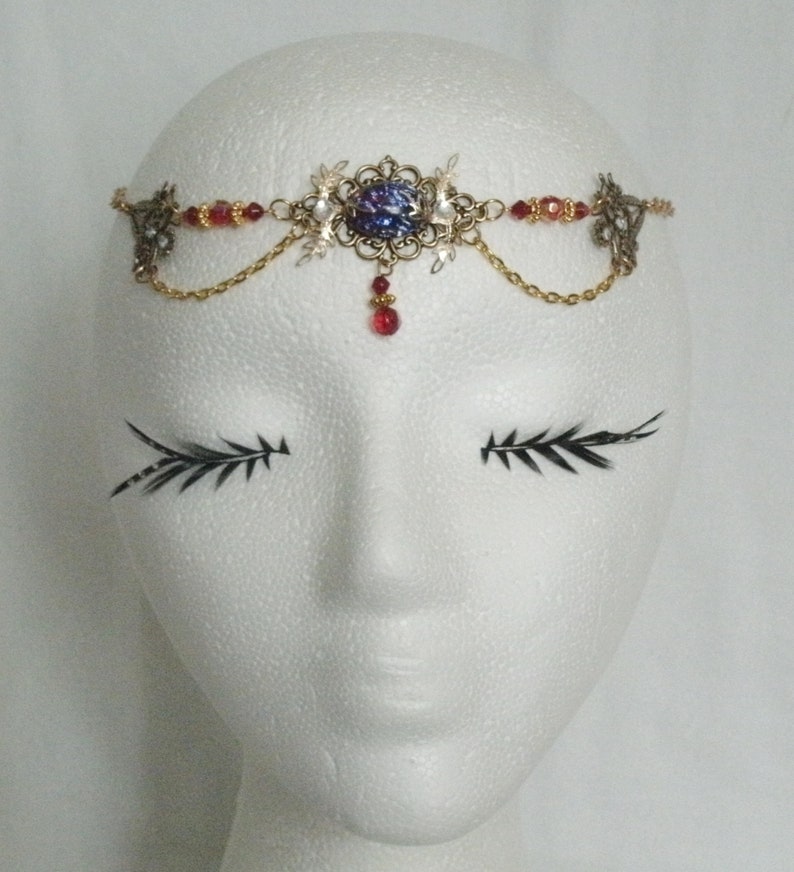 Op de afbeelding: Een gouden en zilveren hoofdband met een grote, iriserende blauwe edelsteen in het midden. De hoofdband is versierd met kleine rode kralen en witte kristallen.