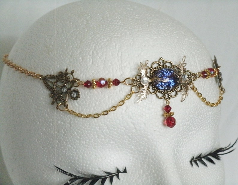 Op de afbeelding: Een gouden en bronzen hoofdtooi met een blauwe, iriserende edelsteen, rode kralen en een ketting. De hoofdtooi is op een wit mannequinhoofd.