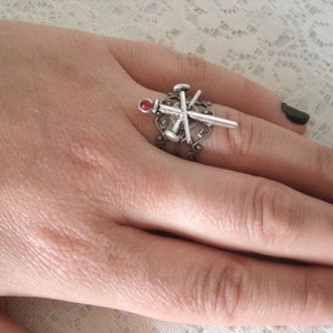 May include: A silver ring with a red stone and a design of crossed nails. The ring is worn on a finger with black nail polish.