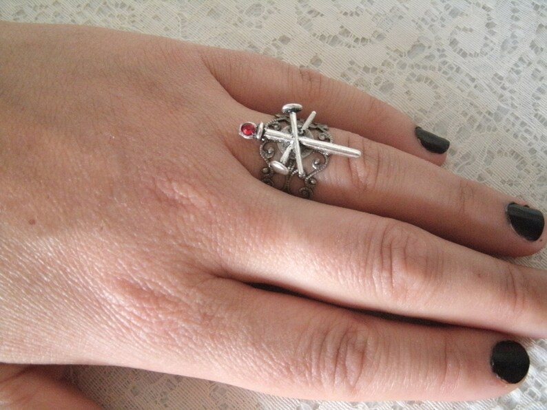 May include: A silver ring with a red stone and a design of crossed nails. The ring is worn on a finger with black nail polish.
