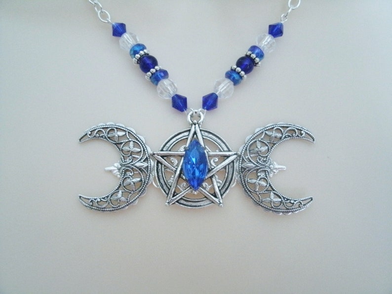 May include: A silver necklace featuring a triple moon pendant with a blue crystal at the centre of a pentagram. The necklace is made with blue and clear glass beads.