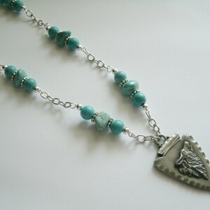 May include: A silver chain necklace with turquoise beads and a silver pendant featuring a wolf's head in a triangular shape.