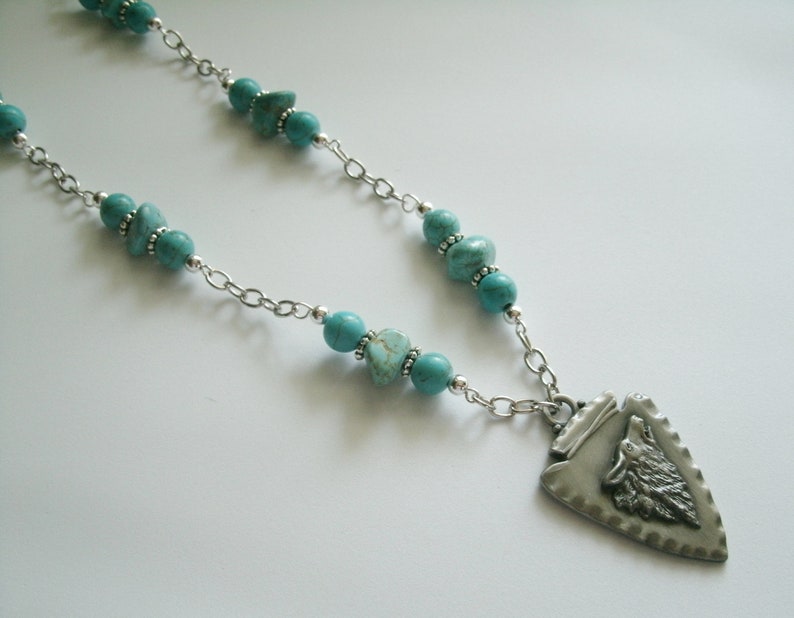 May include: A silver chain necklace with turquoise beads and a silver pendant featuring a wolf's head in a triangular shape.