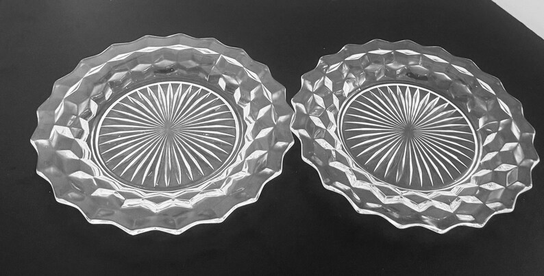 K&ouml;nnte beinhalten: Zwei klare Glasplatten mit einem rautenf&ouml;rmigen Muster an den R&auml;ndern und einem Sonnenstrahl-Design in der Mitte. Die Teller haben einen gewellten Rand und sind zum Servieren von Speisen oder zur Pr&auml;sentation geeignet.