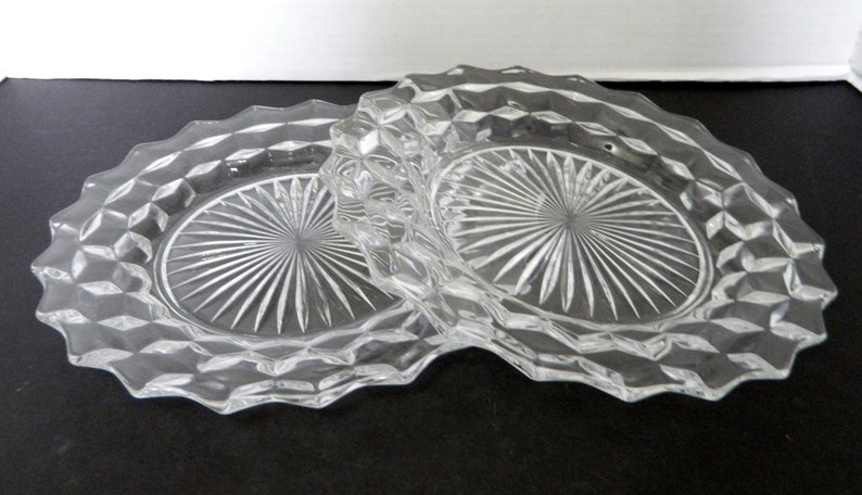 K&ouml;nnte beinhalten: Zwei ovale Teller aus klarem Glas. Jeder Teller hat ein Diamantmuster am Rand und ein Sonnenstrahl-Design in der Mitte. Die Teller bestehen aus klarem Glas und liegen auf einer dunklen Oberfl&auml;che.