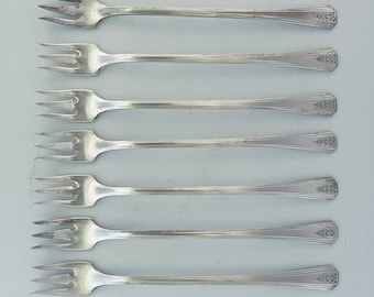 Op de afbeelding: Zeven zilverkleurige vorken met elk drie tanden, horizontaal gerangschikt. De handvatten hebben aan het uiteinde een decoratief bladerpatroon. De vorken staan op een lichtgrijze achtergrond.
