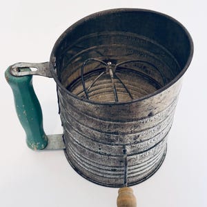 Puede incluir: Colador de harina de metal vintage con mango verde y pomo de madera. El colador tiene medidas para 2, 3, 4 y 5 tazas. El interior presenta un mecanismo de batidor de alambre. El metal tiene un aspecto envejecido.