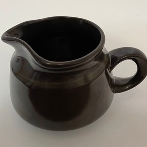 May include: A dark brown ceramic creamer with a rounded body, a pouring spout, and a curved handle. The creamer has a glossy finish and appears to be handmade. This kitchenware item is suitable for serving cream or milk.