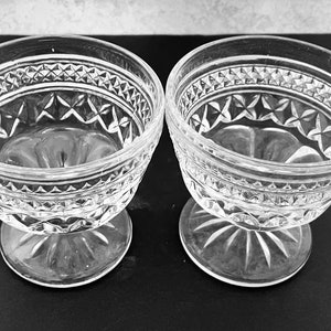 May include: Two clear glass dessert bowls with ornate diamond and star patterns. The bowls have a stemmed base and a decorative rim. The bowls are likely vintage and are suitable for serving ice cream or other desserts.