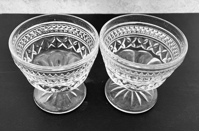 May include: Two clear glass dessert bowls with ornate diamond and star patterns. The bowls have a stemmed base and a decorative rim. The bowls are likely vintage and are suitable for serving ice cream or other desserts.