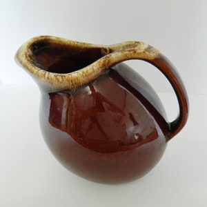 May include: A brown ceramic pitcher with a rounded body and a curved handle. The rim and handle have a lighter, tan-colored trim. The pitcher has a glossy finish, reflecting light. This vintage-style pitcher is suitable for serving beverages.
