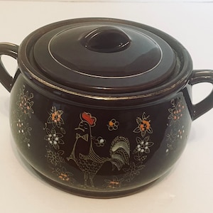 Puede incluir: Una cazuela de cerámica marrón oscuro con tapa, con un diseño de gallo y flores. El plato tiene dos asas y un borde dorado. El gallo es negro con cresta roja y las flores son naranjas y blancas.
